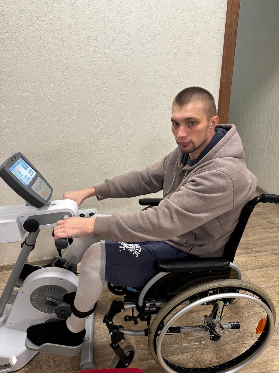 Александр в инвалидной коляске занимается на механическом велотренажёре в реабилитационном помещении, видна лечебная тренировка и адаптация.