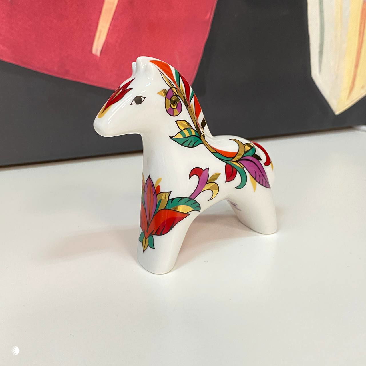 Небольшая белая керамическая фигурка лошадки с ярким растительным орнаментом, снятая на светлом столе, предмет сувенирной эстетики.