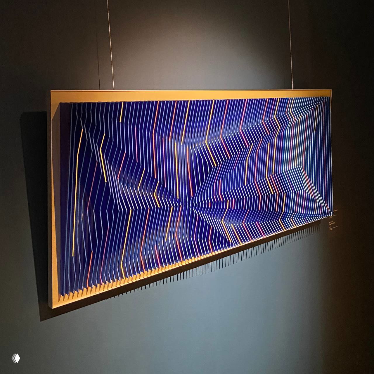 Инсталляция или картина с оптическим ритмом: синие полосы и объёмная структура на стене, современный арт-объект в затемнённой комнате.