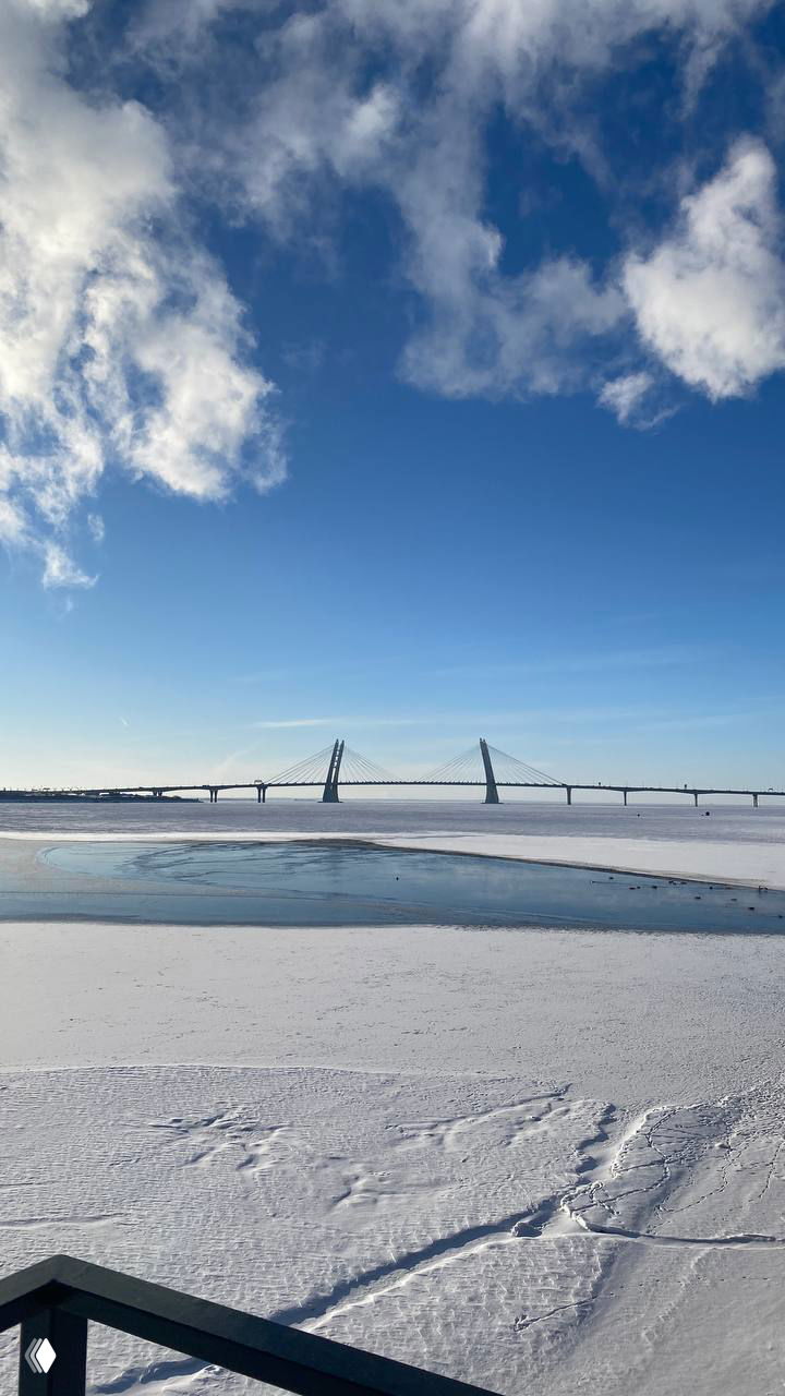Заливной зимний пейзаж с видом на замёрзшую воду и мост на далёком плане, голубое небо и снежная поверхность, холодная городская панорама.