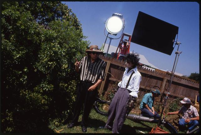 Внешняя съёмочная площадка: персонаж в костюме стоит под дневным светом на фоне техники и членов команды, видно осветительные рамы.