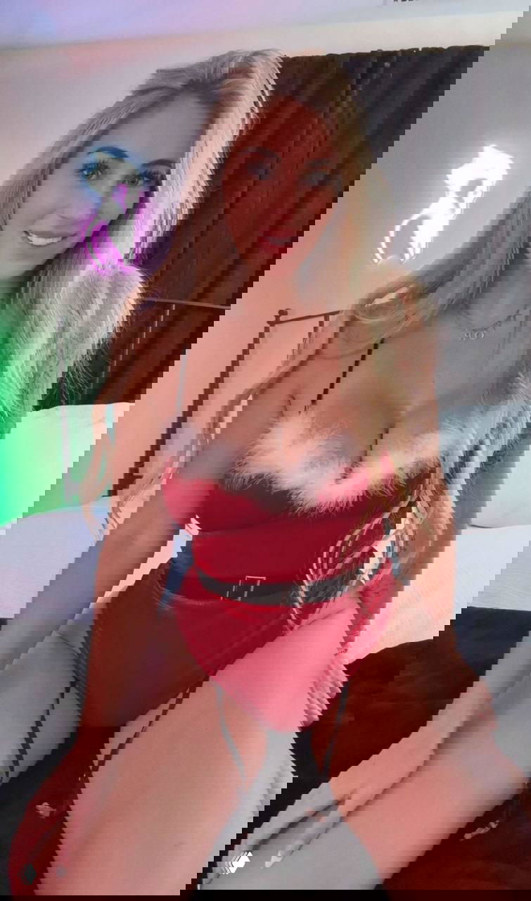 Модель в праздничном красном белье сидит на кровати в студийном интерьере с неоновым акцентом на стене, кадр подчёркивает фигуру и позу.