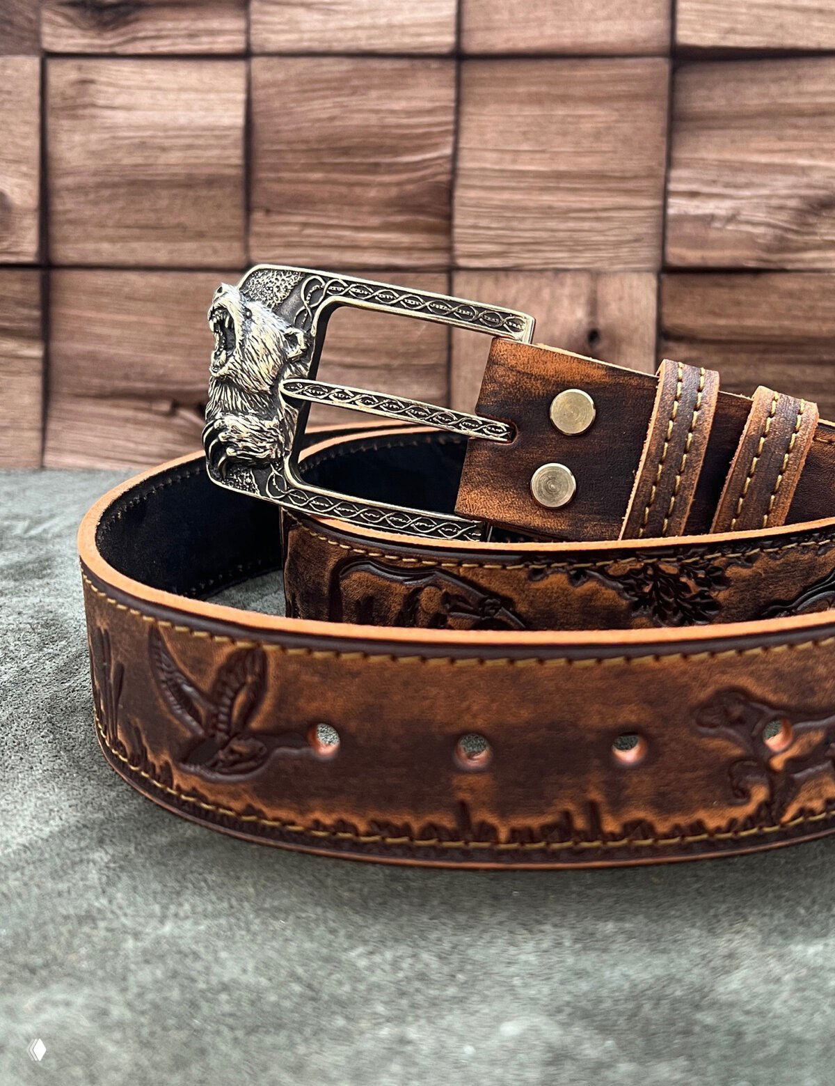 Бежево-коричневый кожаный ремень с металлизованной пряжкой в виде головы медведя и рельефным витиеватым рисунком на коже.