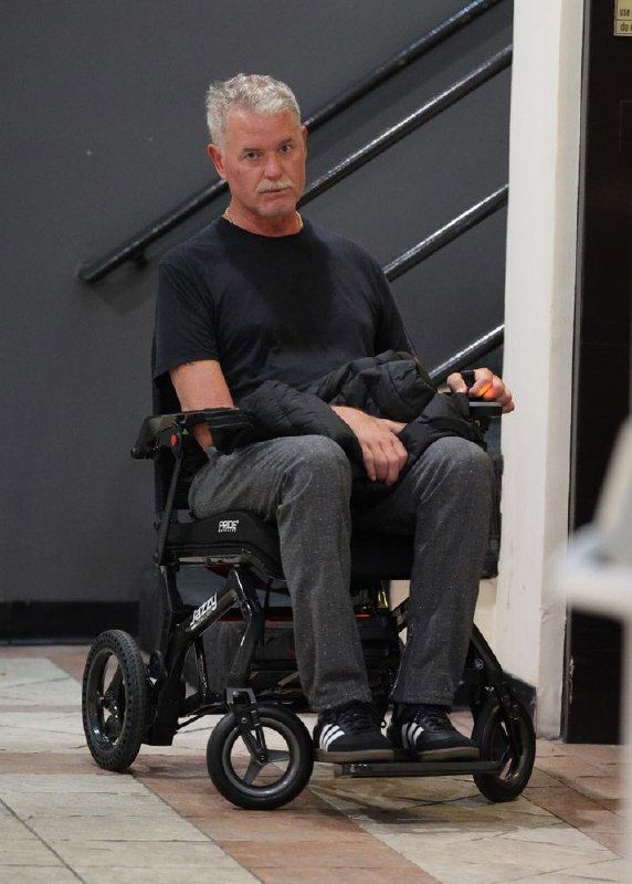 Мужчина средних‑пожилых лет, сидящий в электрическом инвалидном кресле в здании; видно колёса, одежду и общий вид в полный рост.