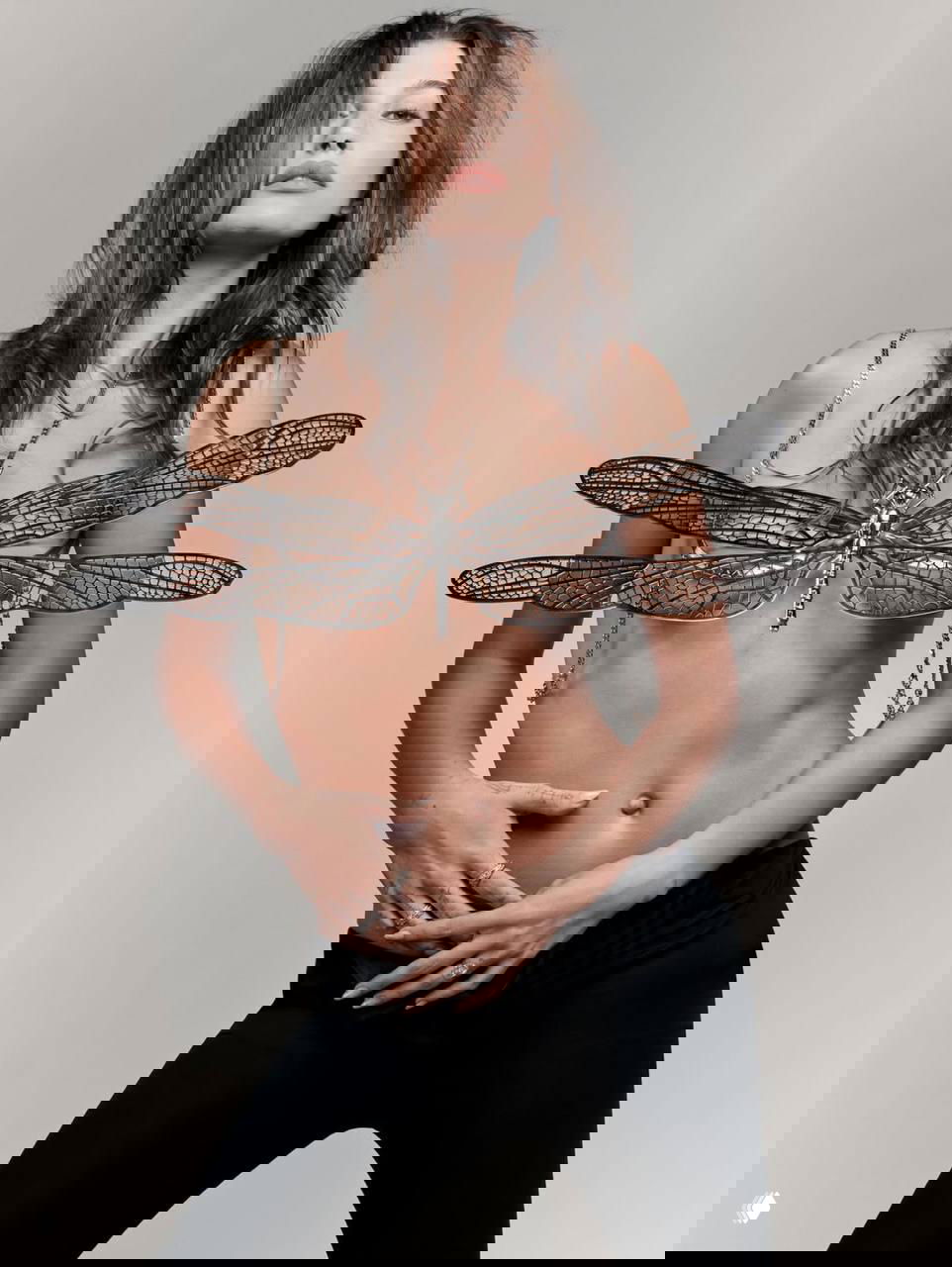 Кадр Хейли Бибер в нейтральных тонах с декоративной брошей‑стрекозой на груди; полуобнажённый модный образ с акцентом на торсе.