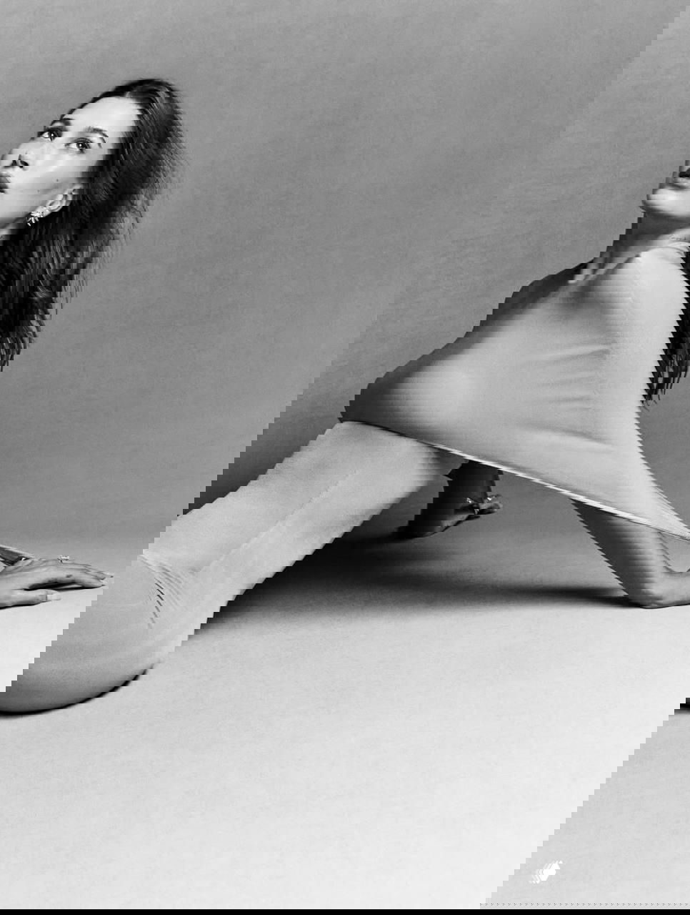 Чёрно‑белый снимок Хейли Бибер в сидячей позе в обтягивающем платье, вытянутая рука и подчёркнутые изгибы силуэта на студийном фоне.