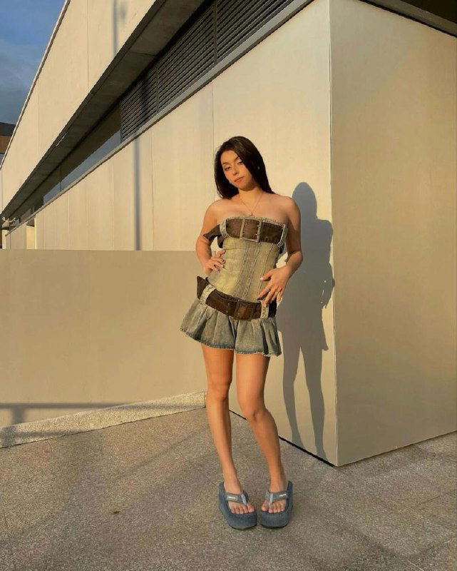 Молодая женщина в коротком платье стоит на терассе при заходе солнца, длинные ноги и расслабленная поза на урбанистическом фоне.