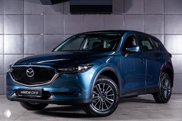 Синий Mazda CX-5 на выставочном стенде в студийной подсветке: кроссовер показан под углом, видна решётка радиатора и дизайн колёсных дисков.