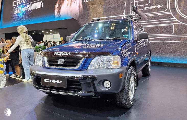 Синий Honda CR-V предыдущего поколения на автошоу: автомобиль представлен спереди, заметна эмблема и защита бампера, окружение выставки и посетители.