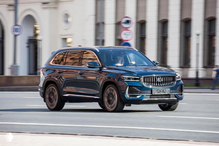 Тёмно-серый Geely Monjaro в городской среде: кроссовер снят крупным планом на дорогe у зданий, видна форма кузова и колёсные диски в движении.