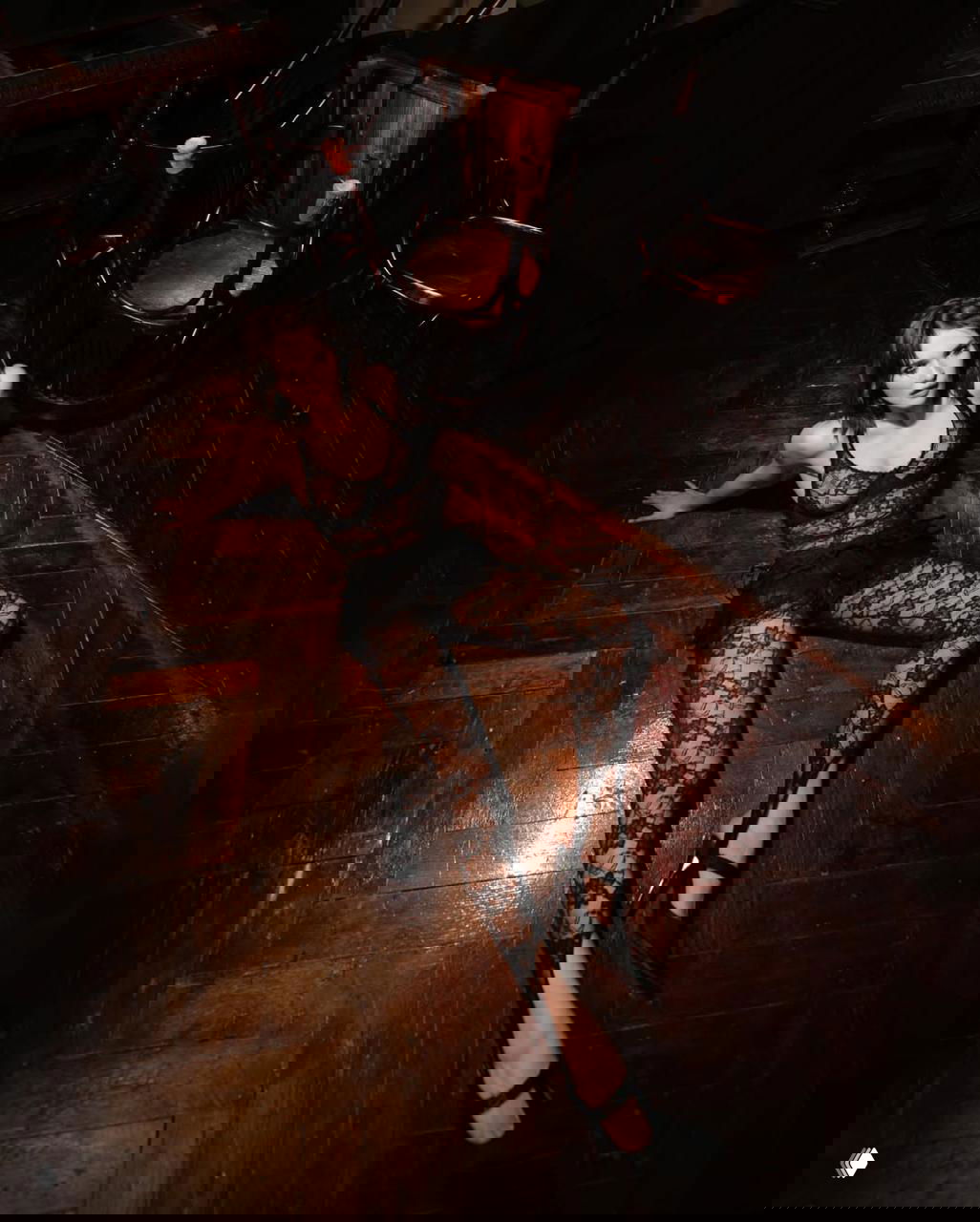 Модель в кружевном цельном наряде на деревянном полу студии; кадр сверху, длинные ноги в туфлях и винтажная мебель на заднем плане создают ретро-настроение.