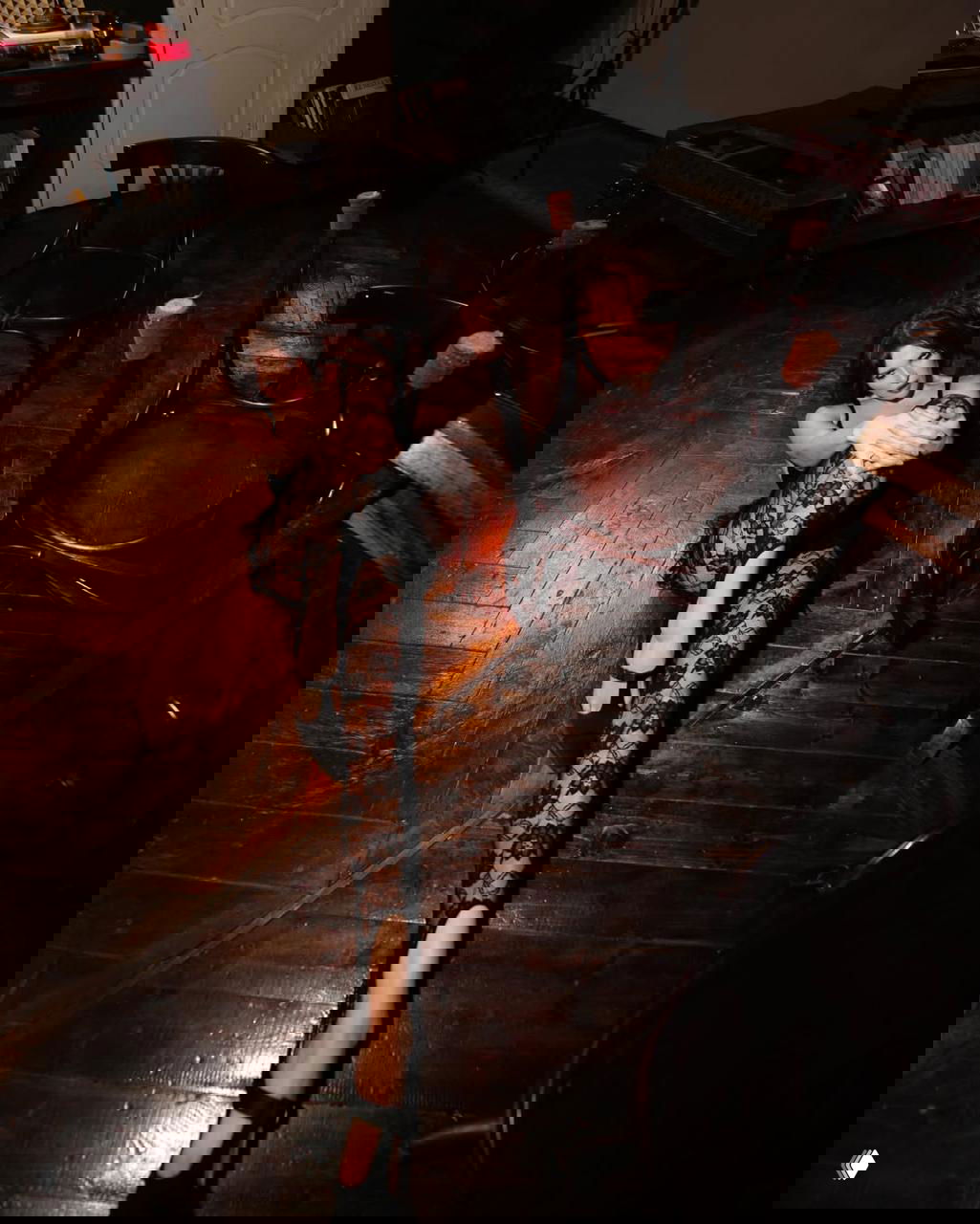 Сидящая поза модели в кружевном комбинезоне на тёмном деревянном полу; рядом перевёрнутые стулья, сцена выглядит как интимная студийная композиция.