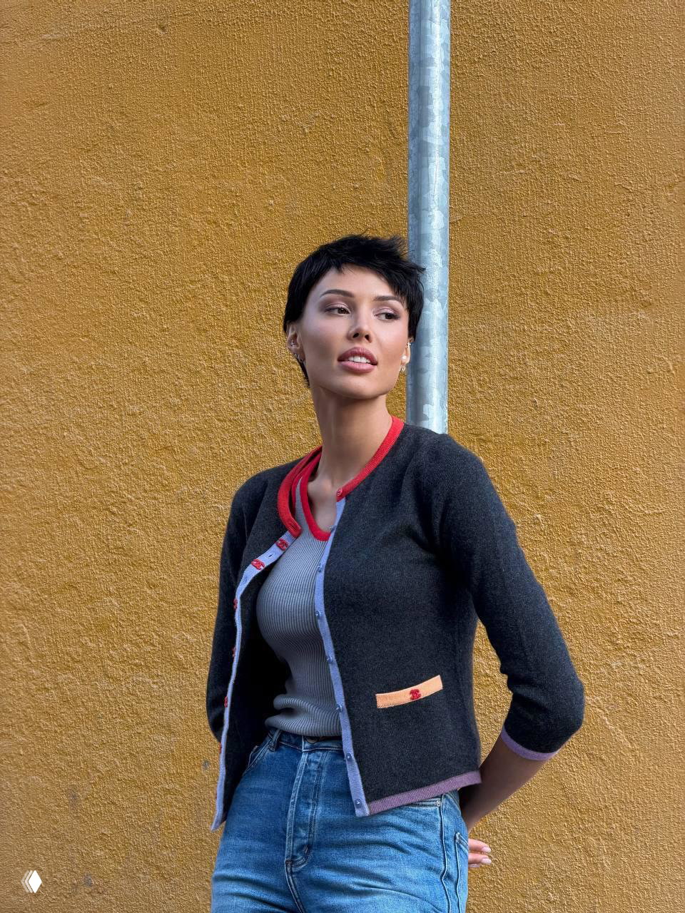 Женщина с короткой тёмной стрижкой в кардигане и джинсах позирует у жёлтой стены — уличный кадр, демонстрирующий новый образ.