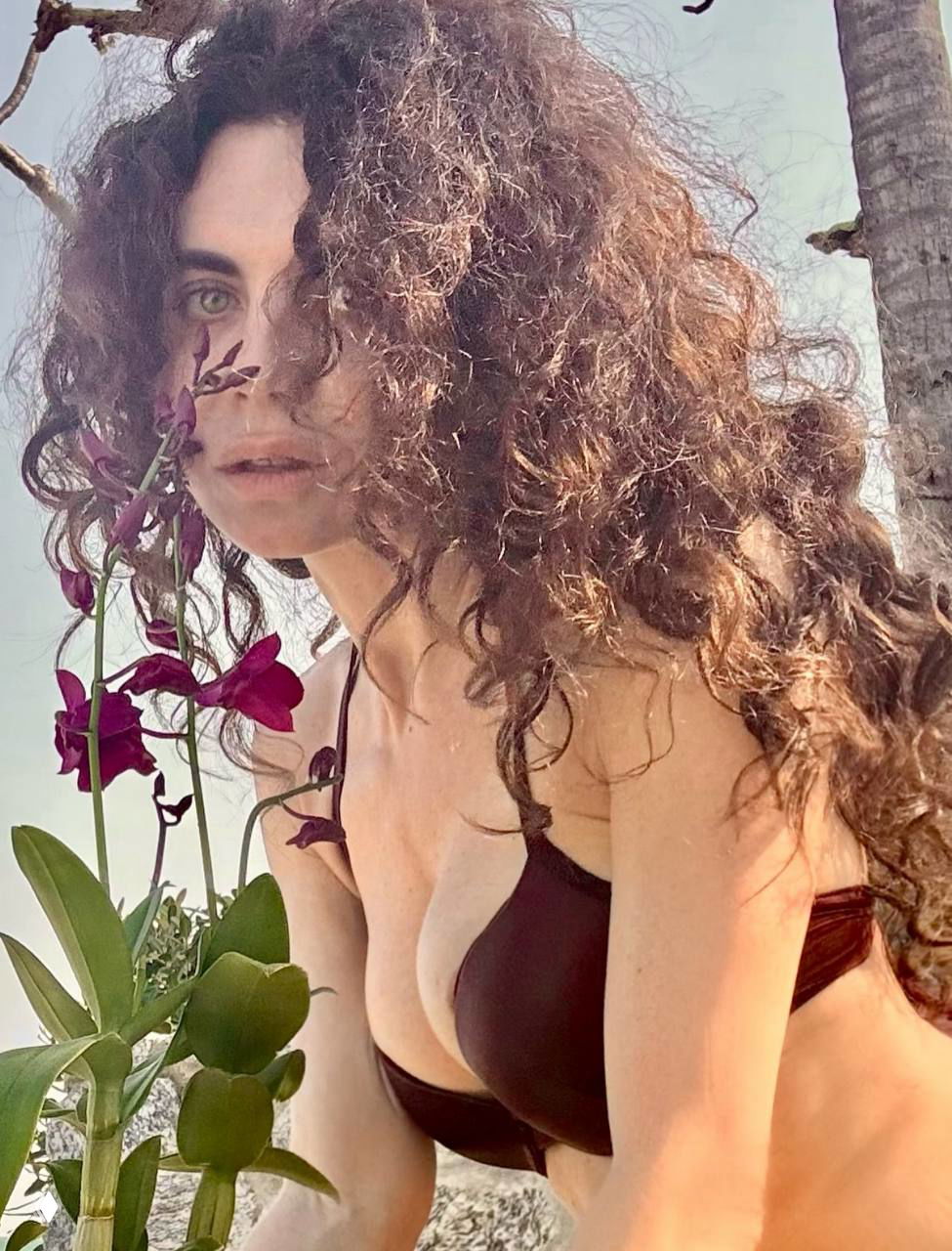 Крупный план Ани Плетневой в коричневом купальнике у орхидей: естественный образ, распущенные кудри и пляжный фон виднеются позади