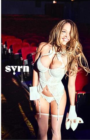 Модель в светлом кружевном корсете и трусиках с подтяжками, позирует сидя в кинозале; заметен логотип бренда и ретро‑стилизация.