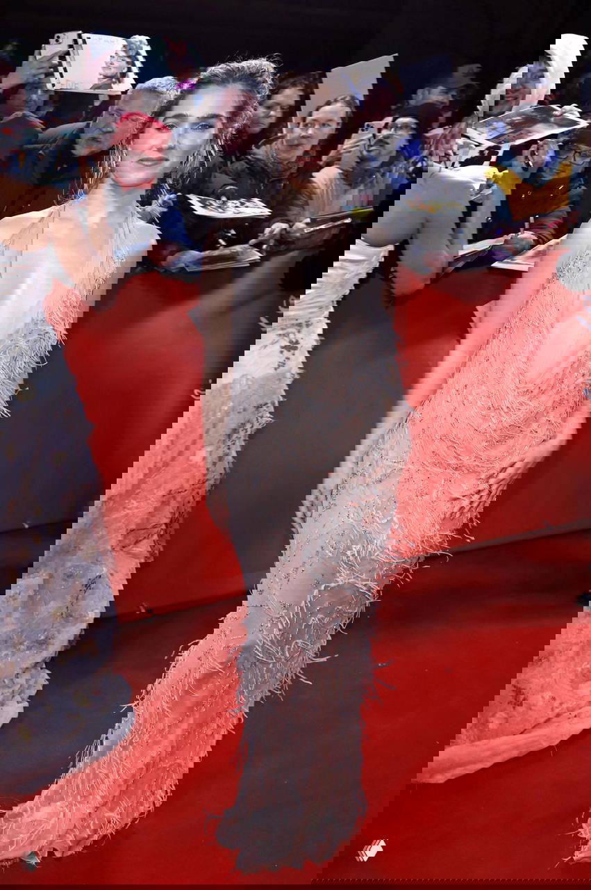 Молодая женщина в светло-розовом платье с глубоким декольте на красной дорожке, зрители и папарацци позади барьера.