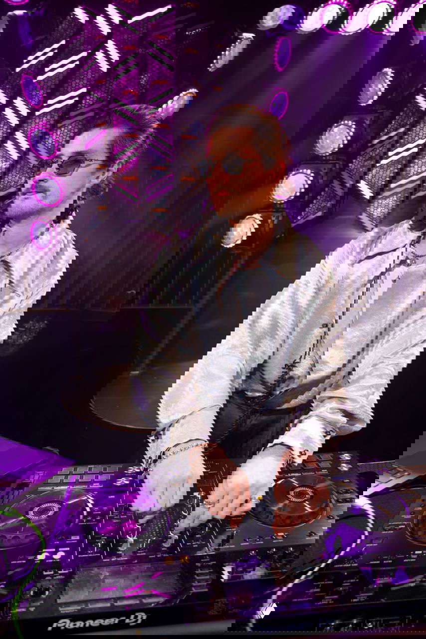 Ди-джей за пультом в переливающейся рубашке управляет сетом на фоне подсветки и круговых светильников вечеринки в клубном пространств