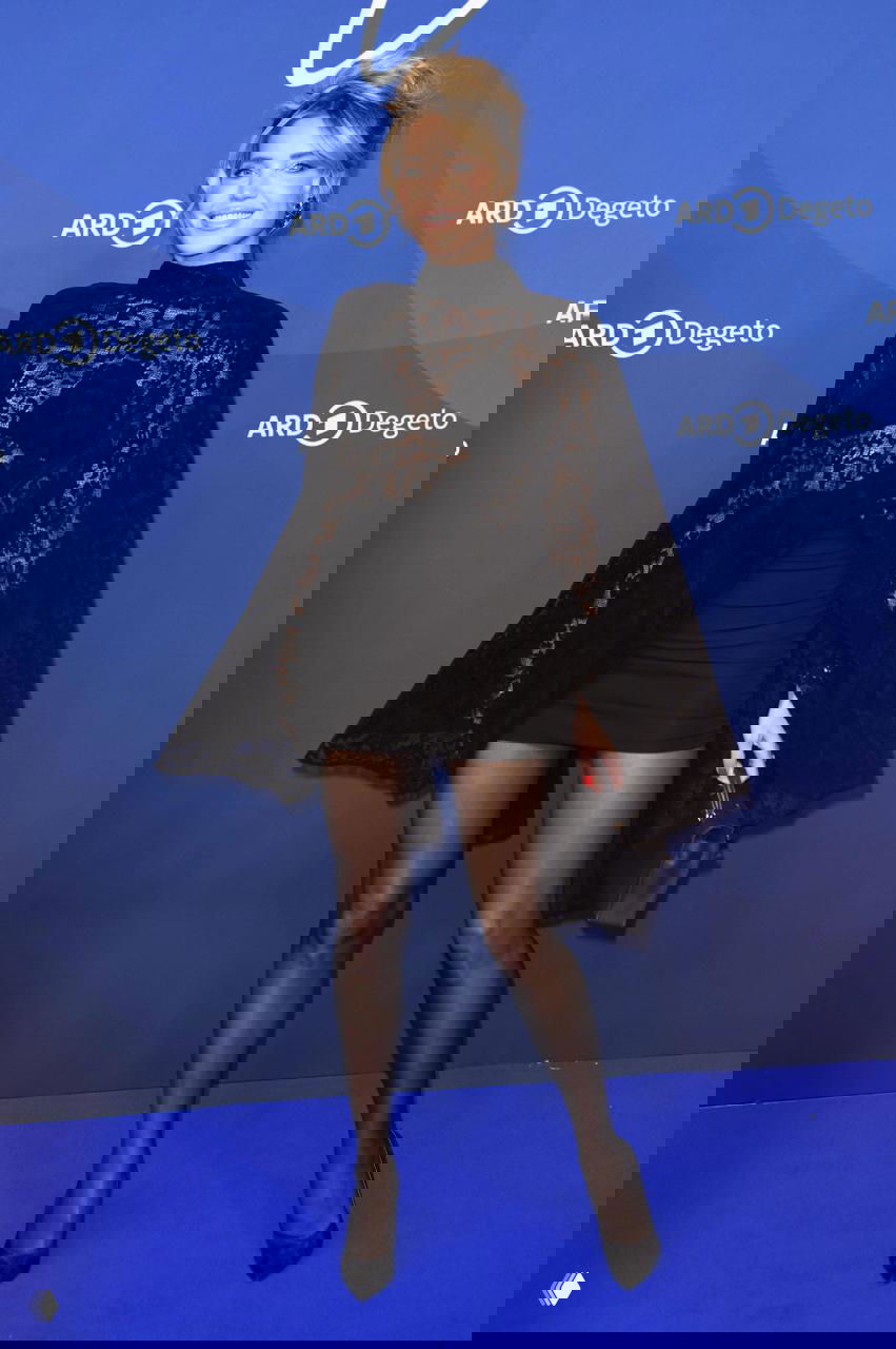 Женщина в черном кружевном плаще и коротком платье позирует на синем фоне промо-ивента, высокий рост и тонкие чулки.