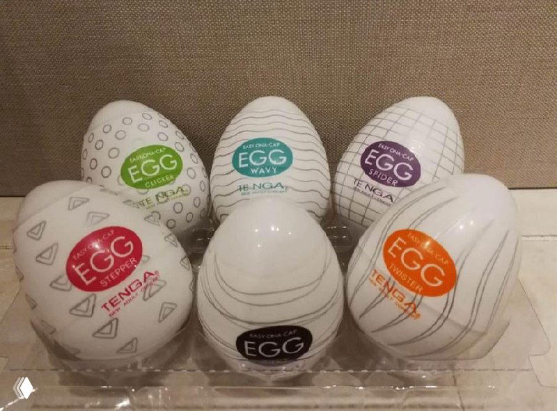 Несколько упаковок и устройств бренда Tenga на витрине/полке; изображение с артефактами, демонстрирующее товарную линейку производителя.