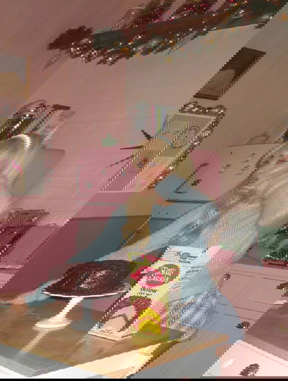 Женщина в ретро-розовой кухне позирует рядом с тортом и коробкой для выпечки, кадр передаёт уютную винтажную атмосферу праздника.