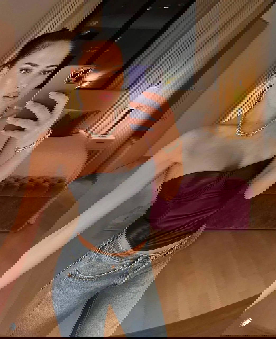 Женщина делает зеркальное селфи в спальне в черном корсете и джинсах, в кадре видна кровать и ночник за ней.