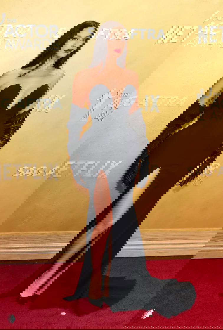 Женщина в облегающем черном платье с глубоким декольте и красной помадой стоит на красной дорожке рядом с логотипами премии и позирует фотографам.