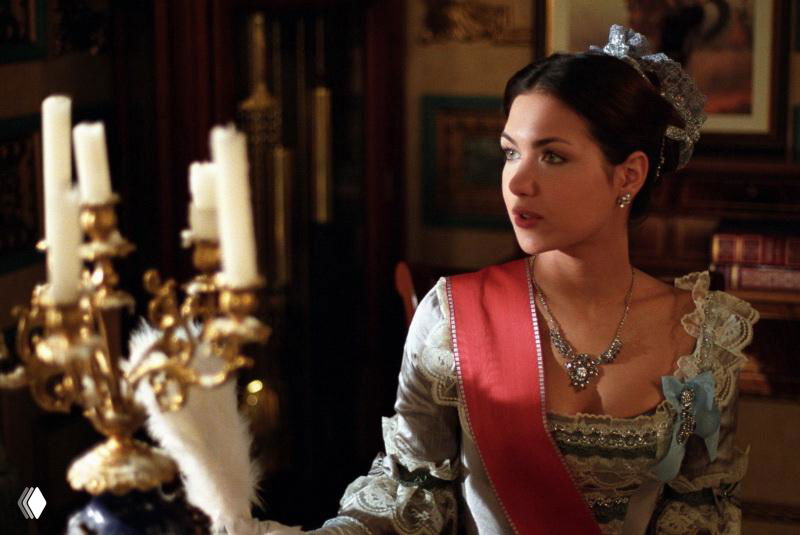 Кадр в антураже дворца: женщина в историческом платье и украшениях возле подсвечника; сцена из костюмного телесериала.