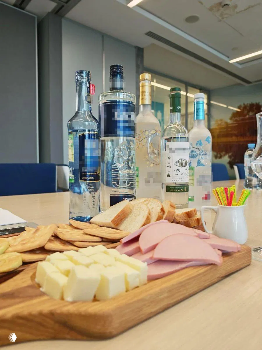 Несколько бутылок водки на столе в офисном интерьере и разделочная доска с хлебом, сыром и колбасой — подготовка дегустации и сопутствующие закуски.