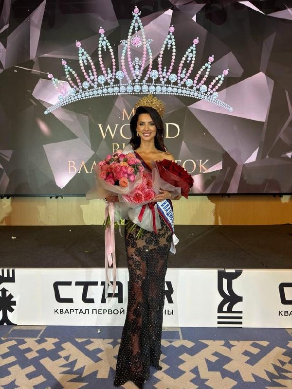 Женщина в короне и с большим букетом стоит на сцене финала конкурса красоты на фоне декоративного изображения короны и экрана.