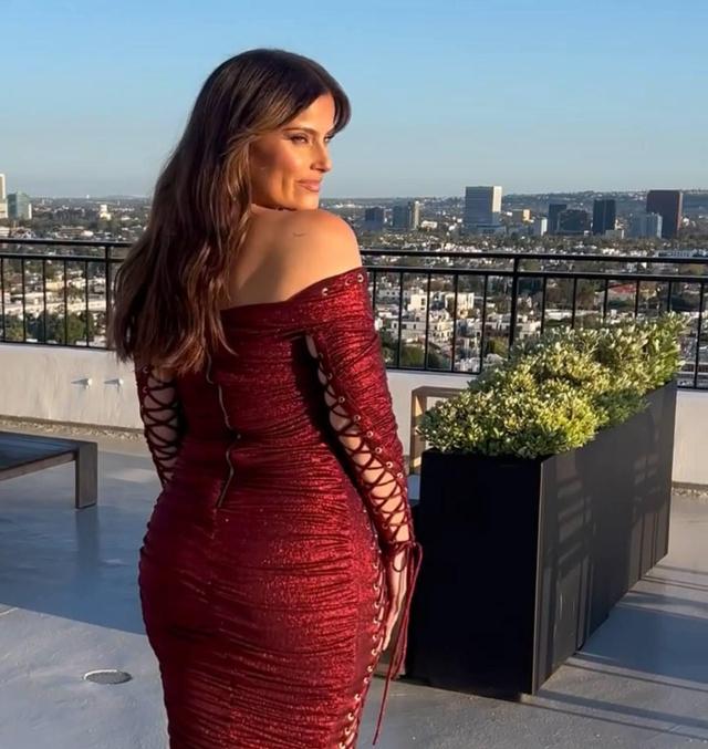 Женщина в приталенном бордовом вечернем платье стоит на балконе с видом на город, акцент на силуэте и отделке платья, дневное освещение и урбанистический фон.