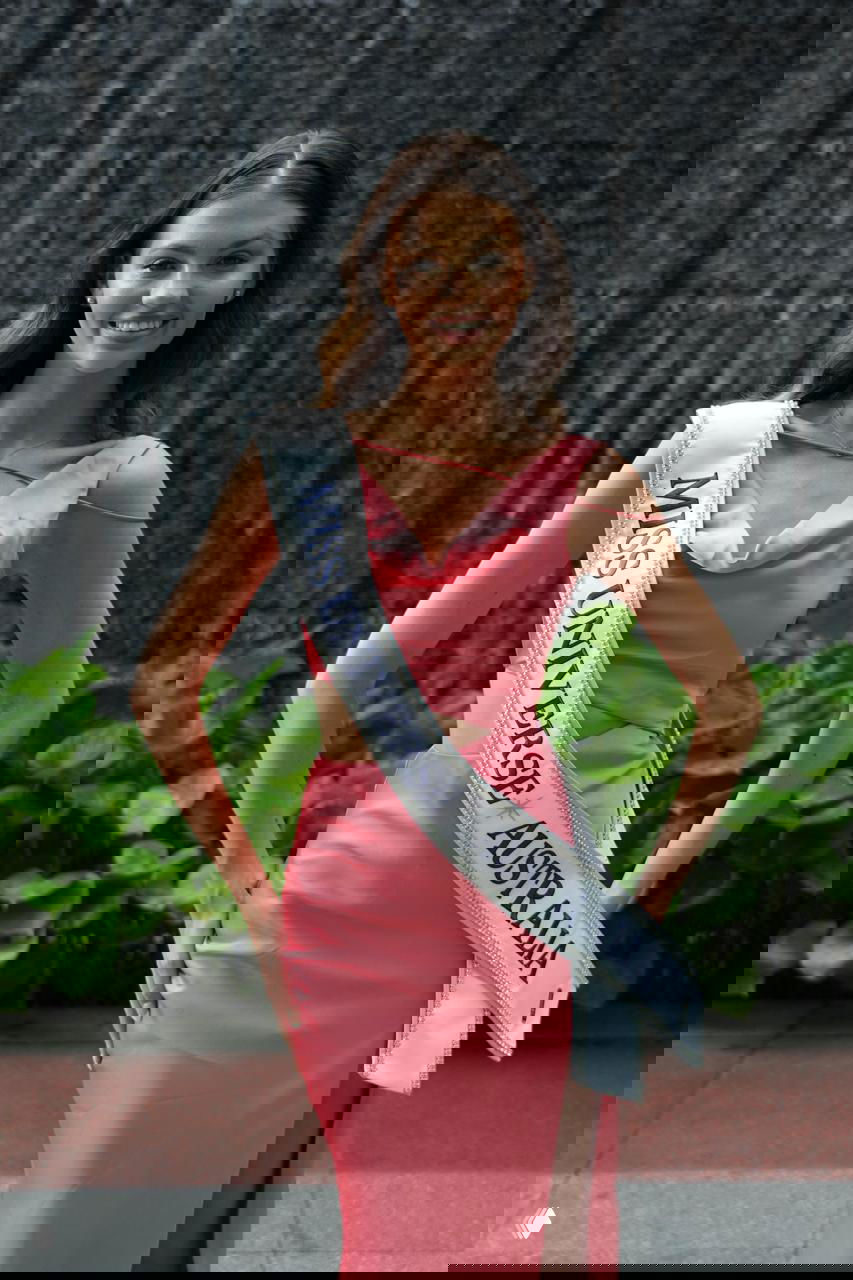 Улыбчивая участница с лентой «MISS UNIVERSE AUSTRALIA» в розовом платье, снимок сделан на фоне зелёной растительности и архитектуры