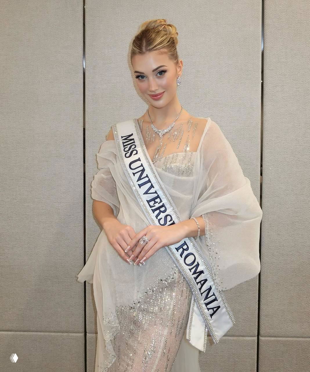 Элегантная участница с лентой «MISS UNIVERSE ROMANIA» в светлом наряде и украшениях, студийный портрет перед серой перегородкой