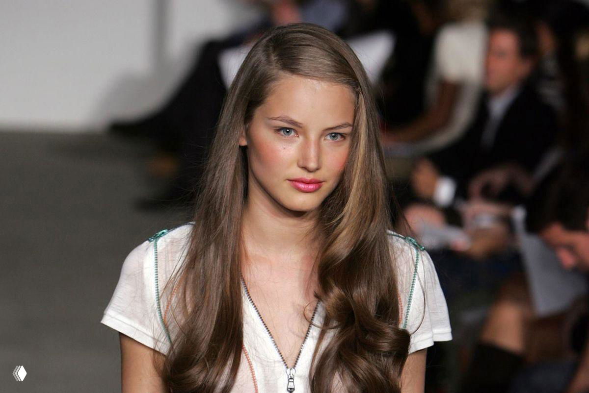 Модель на подиуме: молодая женщина с длинными волосами в естественном макияже во время показа мод, аудитория на заднем плане.