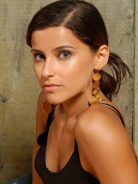 Портрет женщины с зачесанными назад волосами и крупными серьгами‑каплями, нейтральный фон, крупный план лица и плеч, спокойное выражение.