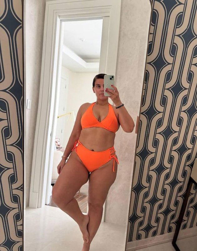 Зеркальное селфи женщины в ярко‑оранжевом бикини в прихожей с узорчатой стеной, полный рост в отражении, в руках виден смартфон и часть интерьера.