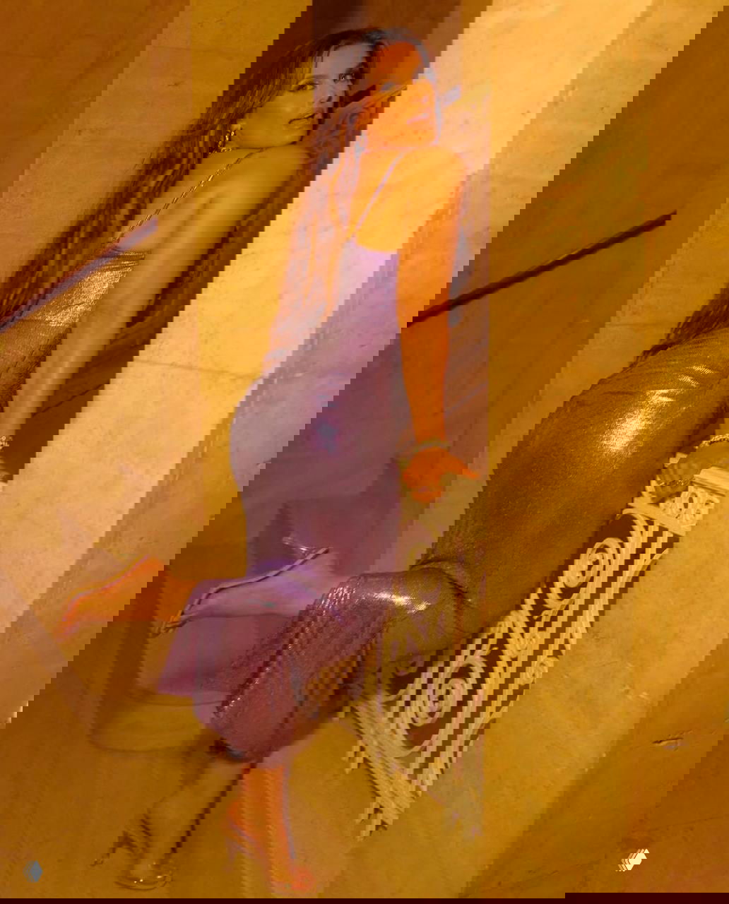 Женщина в блестящем сиреневом платье позирует на лестнице в интерьере с мраморными стенами, тёплое освещение подчёркивает текстуру ткани и изгибы силуэта.