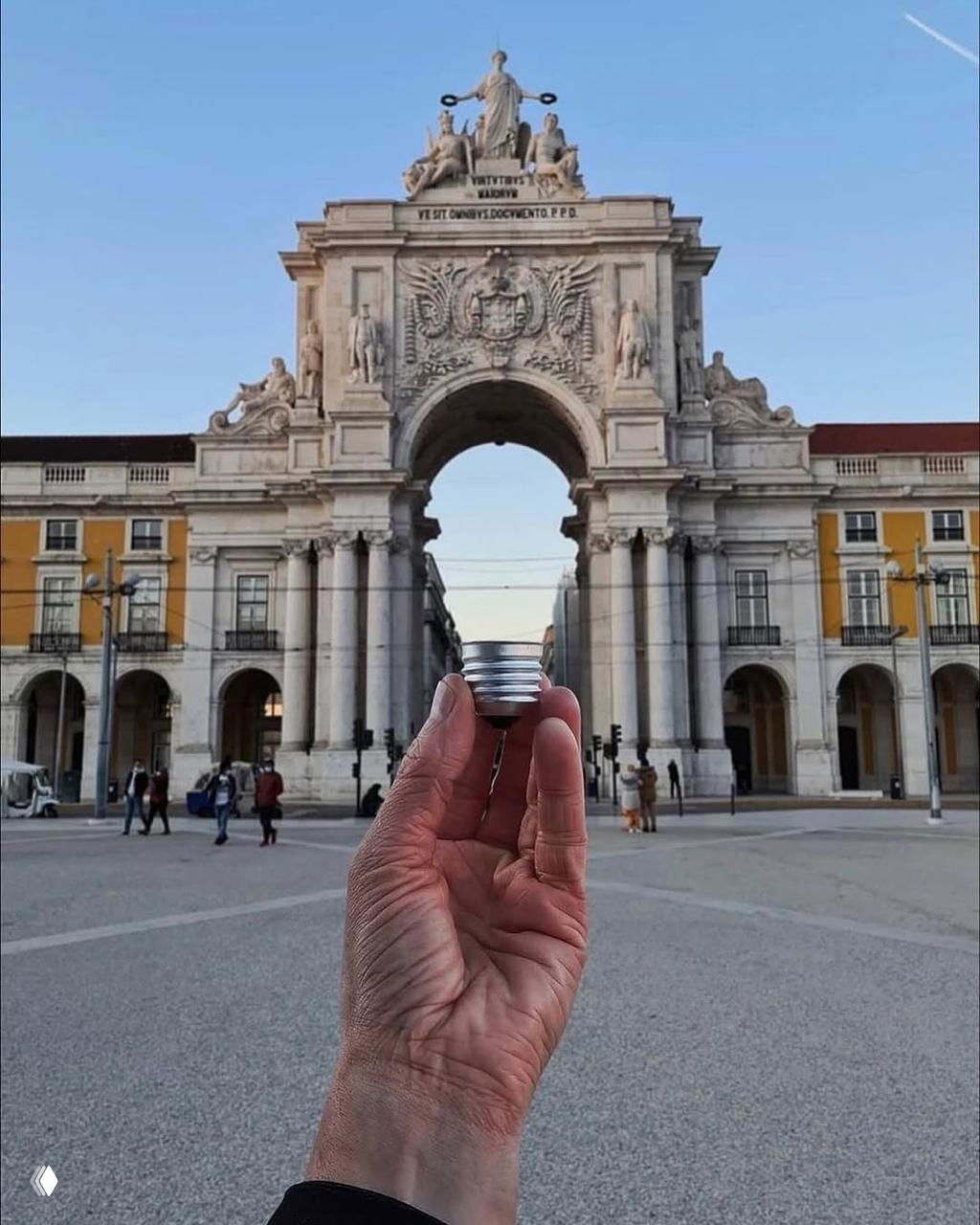 Рука держит небольшой цилиндрический предмет, который в перспективе совпадает с аркой исторического монумента на городской площади.