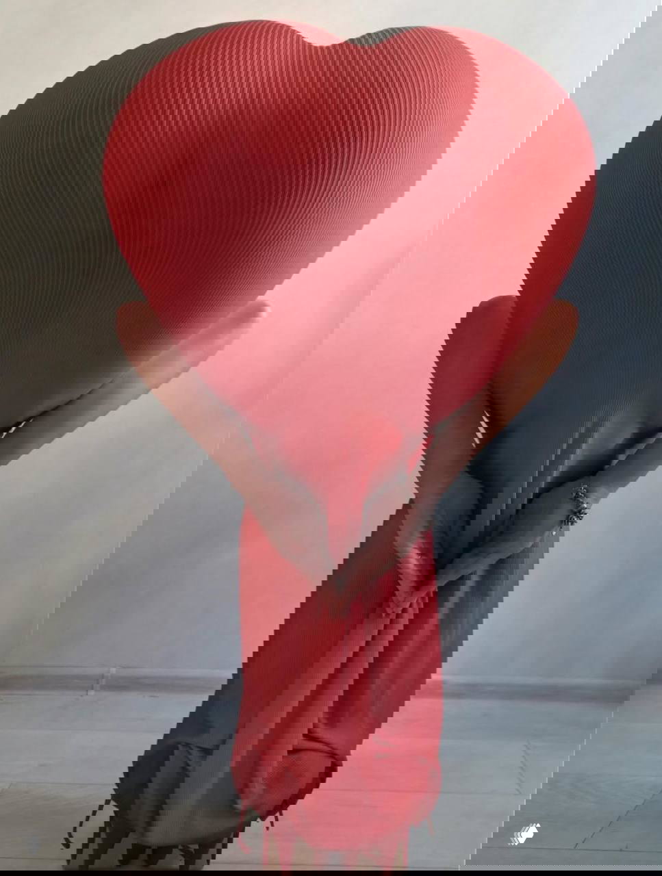 Фигура в красном фактурном платье снята спиной; ткань и поза рук за спиной создают объёмную форму сердца на нейтральном фоне комнаты.