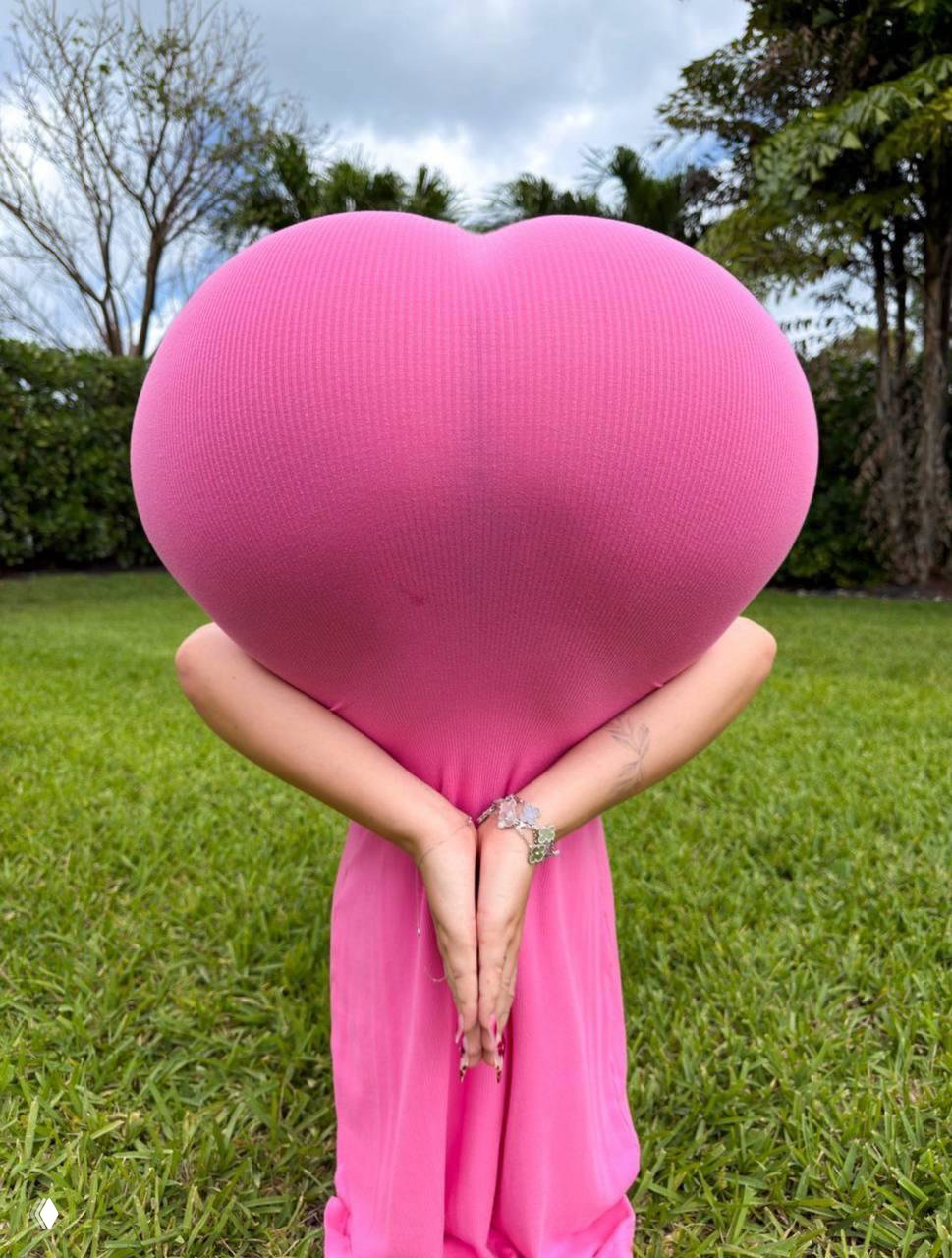 Человек в ярко-розовом платье на фоне зелёного газона; спина к камере, ткань натянута в форму сердца, руки за спиной соединены пальцами внизу.