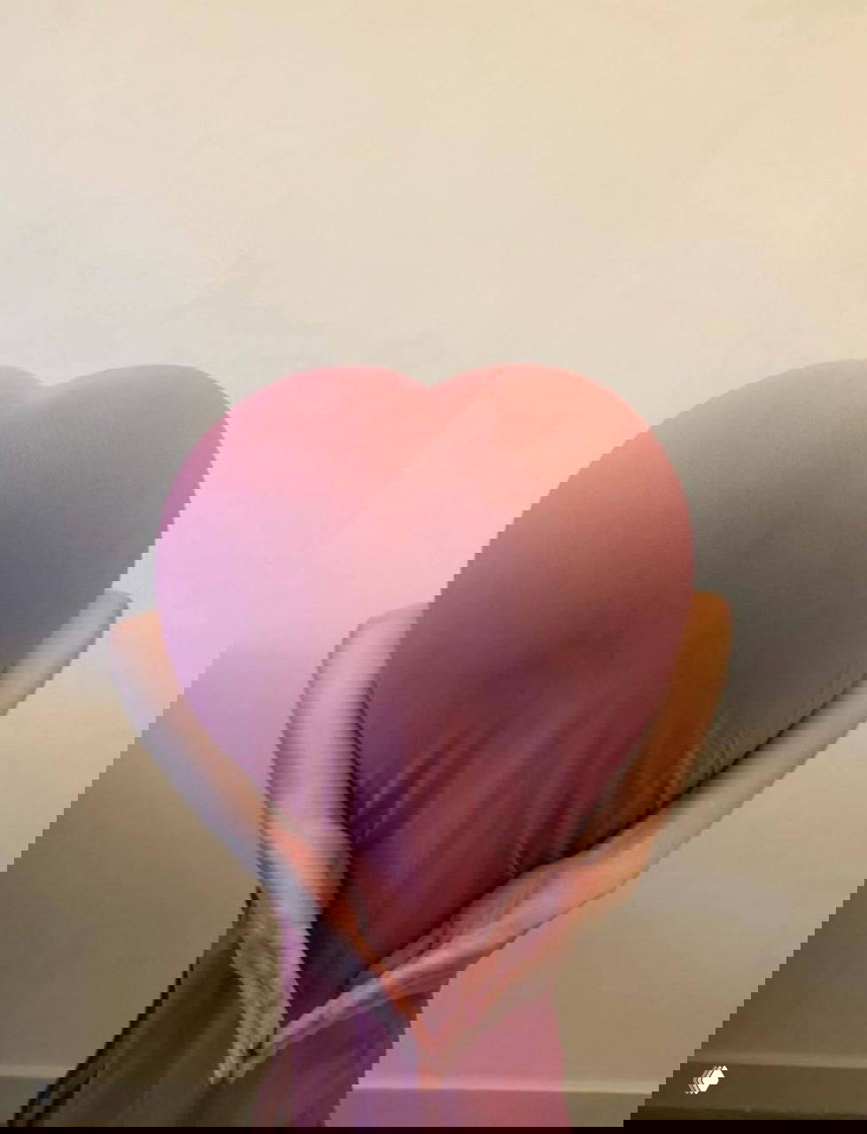 Фигура в пастельном платье спиной к камере; руки за спиной образуют сердечную форму, фон — простая стена, акцент на силуэте и позе.