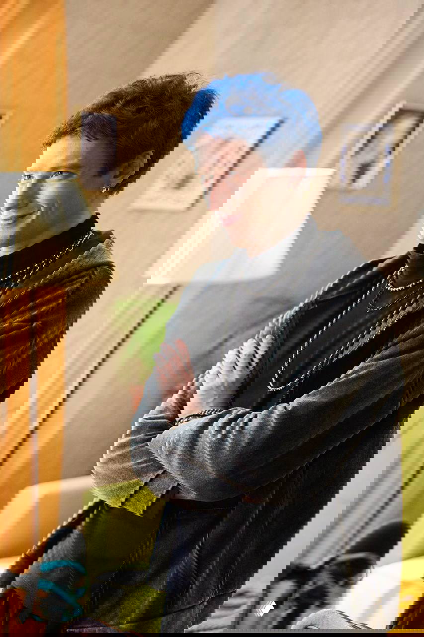 Молодой мужчина с голубыми волосами и украшениями улыбается в гостиной; промо‑кадр шортс‑сериала «Усынови хакера», на переднем плане виден микрофон.