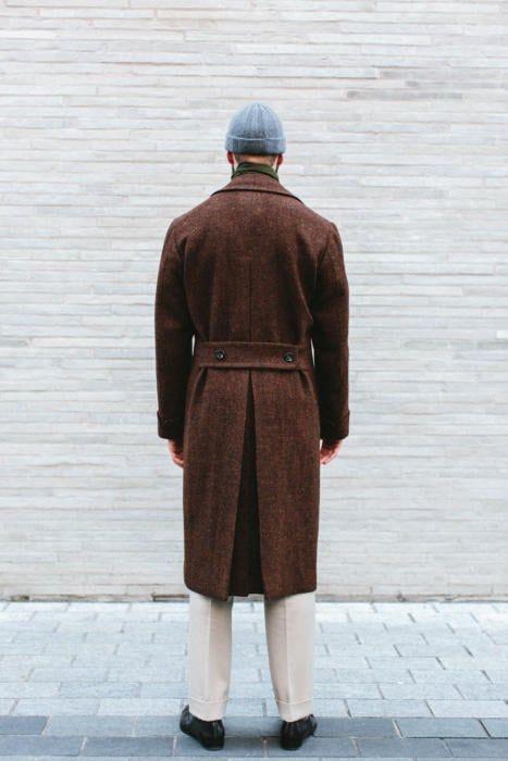 Вид сзади: мужчина в длинном коричневом пальто и серой шапке стоит у светлой кирпичной стены, подчёркнут строгий силуэт и длина изделия.