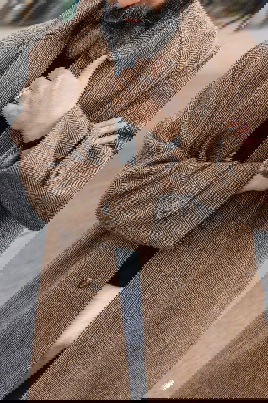 Крупный кадр бородатого мужчины в коричневом твидовом пальто ёлочкой; видна текстура ткани, пуговицы и рука с перстнем, акцент на деталях.