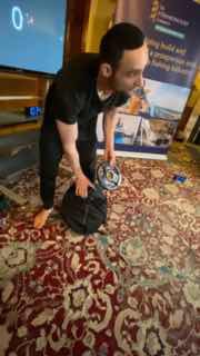 Стоп-кадр: мужчина в чёрной футболке наклоняется над сумкой на узорном ковре в комнате — кадр из видеозаписи тренировки.