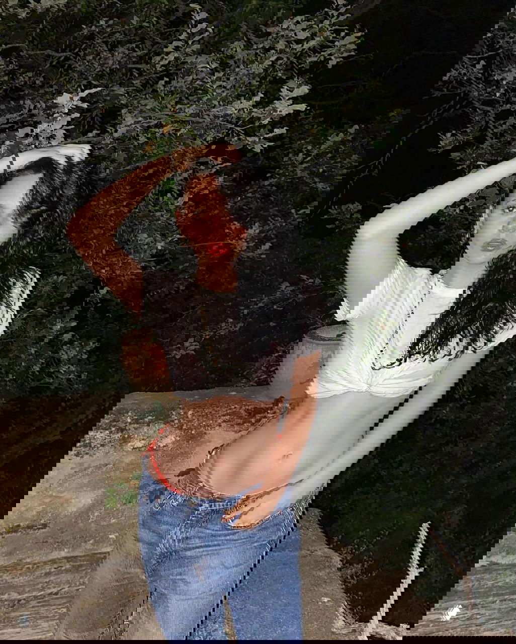 Ночная уличная фотография: молодая женщина в белой футболке и джинсах, поза с рукой в волосах на фоне кустов и каменной кладки.