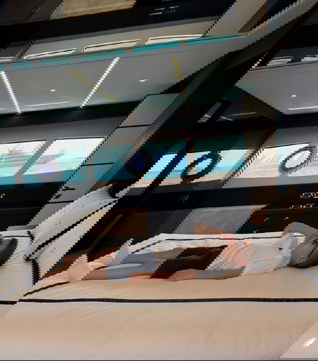 Сын Марк лежит на кровати в каюте яхты в белой футболке и темных шортах; через иллюминаторы виднеется море.