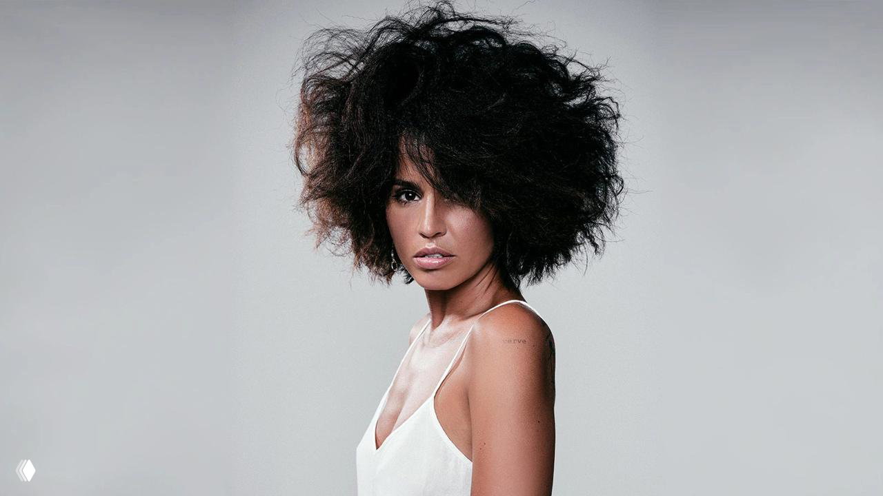 Портретная фотография певицы в светлом образе с объёмной причёской на нейтральном фоне, крупный студийный план в рекламном стиле.