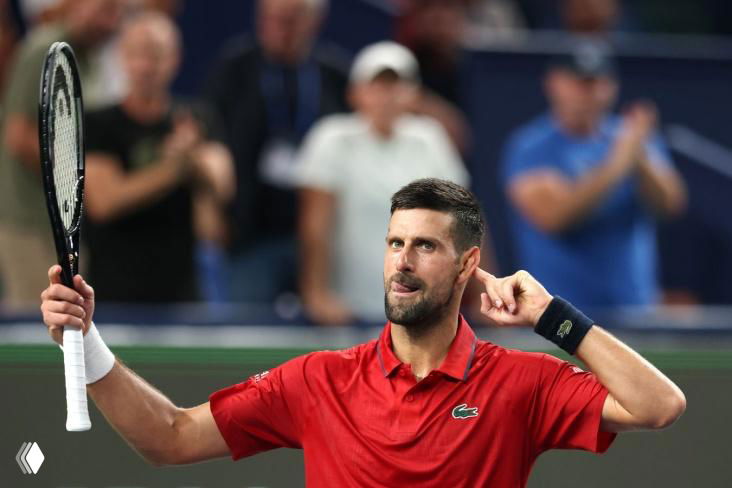 Игрок на теннисном корте в яркой рубашке поднимает ракетку в сторону трибун, публика на заднем фоне, кадр в зале соревнований.