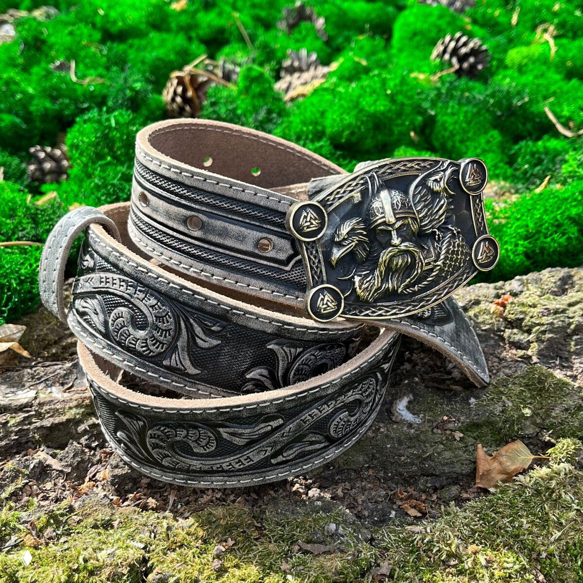 Несколько серых ремней, скрученных в спираль, на фоне зелёного мха; видна крупная металлическая пряжка с барельефом викинга.