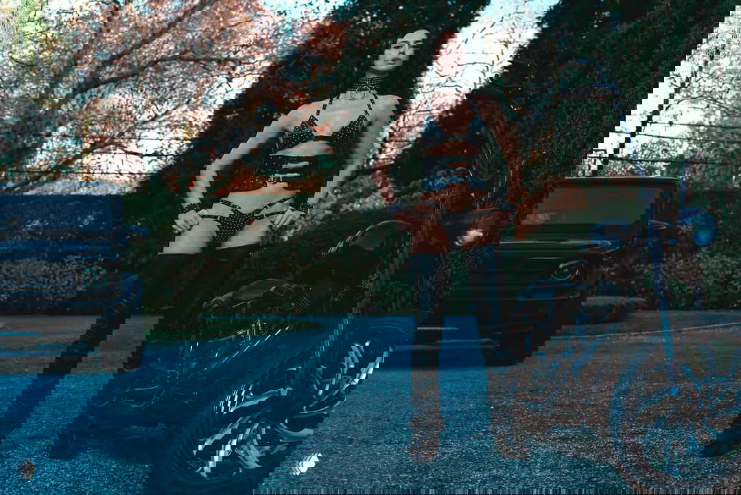 Цветная фотография: женщина в откровенном комплекте и высоких сапогах стоит рядом с мотоциклом на дорожке на фоне деревьев и кустарника.